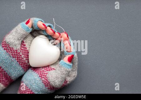 Weibliche Hände im warmen Winter gestrickte Handschuhe mit Mustern, mit mattem Herzen und mit schöner Maniküre - rot glitzernde Nägel auf grauem Hintergrund mit c Stockfoto