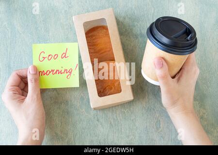 Weibliche Hände halten Notiz mit Worten GUTEN Morgen, Tasse Kaffee, Tastatur, Kaktus und Mause auf einem Holztisch. Konzept für gute Arbeit. Draufsicht auf den Schreibtisch Stockfoto