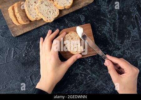 Weibliche Hände halten ein Messer mit Frischkäse und einem Stück Brot auf einem schwarzen Hintergrund. Der Prozess der Herstellung von Sandwiches oder Snacks. Weicher Fokus. Draufsicht. Flach liegend Stockfoto