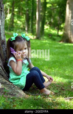 Das kleine Mädchen taucht in das Rollenspiel ein, während sie mit ihrem imaginären Freund am Sprechtelefon chattet. Sie hat Zöpfe und grüne Haarbogen. Stockfoto