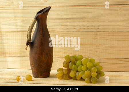 Traubenkrug auf einem natürlichen Holzhintergrund. Rustikaler Retro-Stil. Antiker Tonkrug mit Wein. Grüne saftige reife Trauben der Herbsternte. Das Konzept o Stockfoto