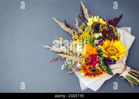 Herbststrauß aus gelb-rot-orangen Blumen, eingewickelt in Papier und auf grauem Hintergrund angeordnet. Sonnenblumen, Amaranth, Gänseblümchen mit Zinnien und Gräsern Stockfoto