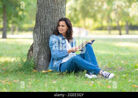 Porträt einer 40-jährigen Frau, die im Park ein Buch liest. Sie macht eine Pause von der Arbeit am Computer und ständig online. Digital Detox Concep Stockfoto