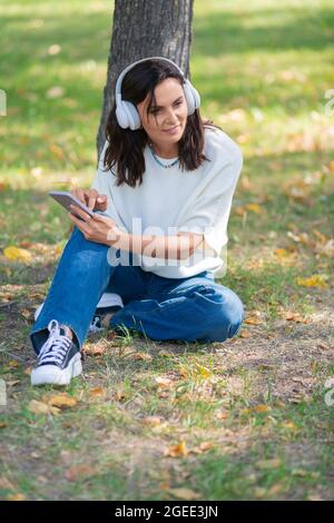Foto einer glücklichen lächelnden 40-jährigen Frau, die Kopfhörer mit einem Smartphone trägt. Sie hört ihre Lieblingsmusik, während sie auf dem Gras im p sitzt Stockfoto