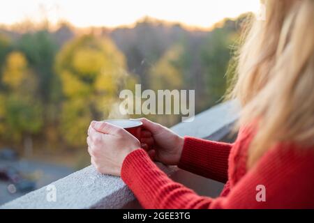 Blonde Frau hält Tasse heißen dampfenden Getränk am Rand des Balkons in hohen Gebäude mit Bäumen und Sonnenuntergang im Hintergrund Stockfoto