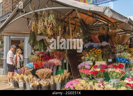 Ein Paar bewundert die bunten Blumen, die an einem Marktstand ausgestellt sind. Blumen werden in Töpfen und Eimern angelegt und ein Geschäft ist der Hintergrund. Stockfoto