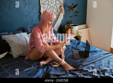Attraktive Frau surft im Internet mit Smartphone, das neben dem Laptop auf dem Bett sitzt Stockfoto