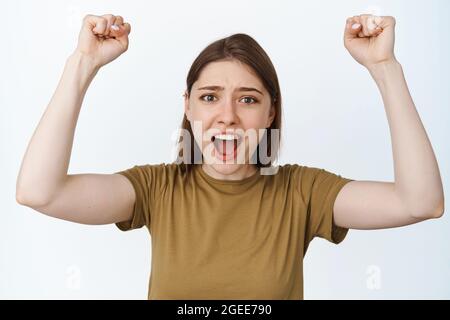 Junge Frau, die singt, die Fäuste hochhebt und schreit, um Sie zu ermutigen, Sportspiele ansieht, für das Team wurzelt, vor weißem Hintergrund steht Stockfoto