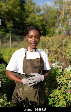 Schwarze Frau mit Tablet lächelnd, während sie in der Nähe von grünen Pflanzen im Garten steht Stockfoto