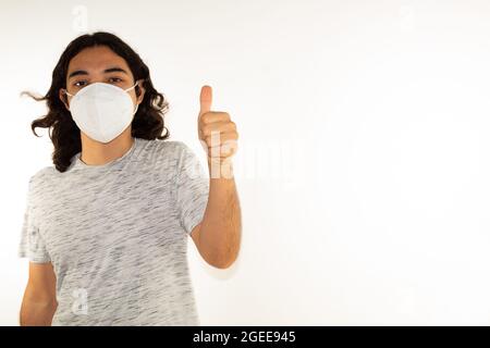 Hispanic Mann mit langen Haaren trägt eine kn95 Gesichtsmaske auf weißem Hintergrund mit einem Daumen nach oben Stockfoto