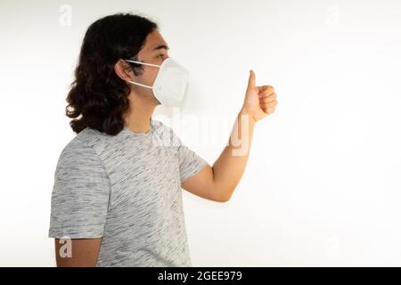 Hispanic Mann mit langen Haaren trägt eine kn95 Gesichtsmaske auf weißem Hintergrund mit einem Daumen nach oben Stockfoto
