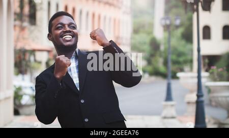 Aufgeregter afrikanischer Geschäftsmann, der die Waffen hochhält und Positivität zum Ausdruck bringt, während er im Freien mit dem Gebäude im Hintergrund steht Stockfoto