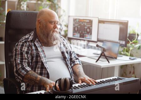 Bärtiger übergewichtiger Mann spielt im Heimstudio neben dem Schreibtisch einen Sound-Synthesizer mit Monitoren Stockfoto