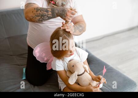 Vater mit Tätowierungen macht Pferdeschwanz zu kleinen Mädchen tragen Feenanzug im Zimmer Stockfoto