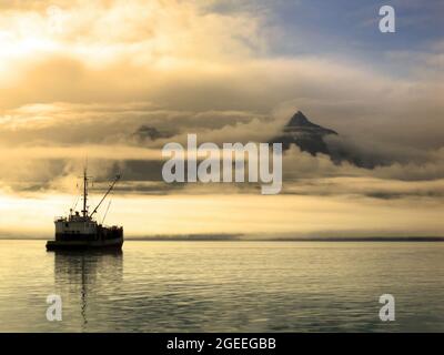 Das Schiff sitzt ruhig an der Bucht von Valdez, während der Sonnenaufgang die Wolken und den Nebel über den Chugach Mountains in Alaska färbt. Der Himmel beginnt sich rechts zu klären Stockfoto