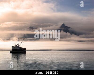 Das Schiff sitzt ruhig an der Bucht von Valdez, während der Sonnenaufgang die Wolken und den Nebel über den Chugach Mountains in Alaska färbt. Himmel klärt sich in der rechten Ecke von i Stockfoto