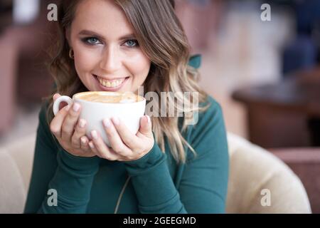 Eine glückliche, schöne Brünette mit einer schönen Frisur, die eine Tasse Kaffee in einem grünen Rollkragen hält, in einem Café sitzt, blickt direkt in die Kamera. Hochwertige Fotos Stockfoto