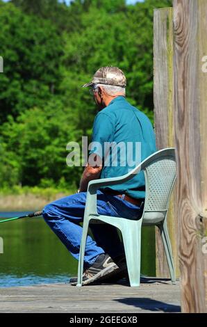 Ein älterer männlicher Fischer sitzt auf einem grünen Stuhl aus Plastik auf einem hölzernen Dock. Er fischt auf einem See nach Brassen. Stockfoto