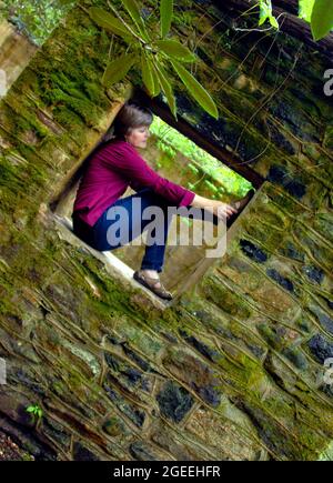 Frau hockt in einem moosbedeckten Fenster, in einem vergessenen Steingebäude. Das Foto ist angewinkelt und sie berührt ihren Schuh. Bild zeigt ein traumhaftes uto Stockfoto
