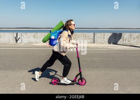 Junge Erwachsene sportliche Hipster Frau im Trainingsanzug, Rucksack und Yogamatte Reiten Kick Scooter schnell gegen konkrete Stadt Flussufer auf Stockfoto