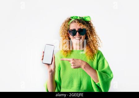 Leuchtend rote lockige Haare lächelnde Frau in herzförmiger Sonnenbrille, sich glücklich fühlend, demonstrierte ihr neues Mobiltelefon und zeigte mit dem Finger auf eine leere Kopie Stockfoto