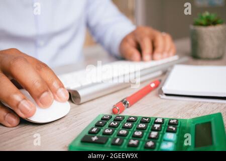 Nahaufnahme der Hände eines hispanischen Mannes, der eine Computermaus mit einem Rechner vor sich benutzt Stockfoto