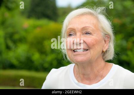 Lächelnde, freundliche 82-jährige Frau in ihrem üppigen grünen Garten, die in einem Nahaufnahme-Porträt von Kopf und Schultern zur Seite schaut Stockfoto