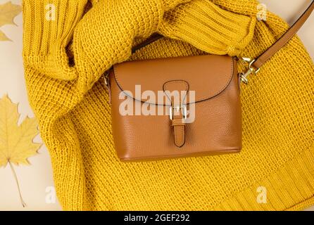 Orangefarbene Ledertasche, Strickpullover, Herbstblätter auf beigefarbenem braunen Hintergrund. Trendige Herbst-Accessoires. Gemütliches Outfit Stockfoto