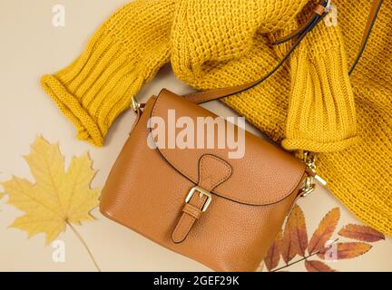 Orangefarbene Ledertasche, Strickpullover, Herbstblätter auf beigefarbenem braunen Hintergrund. Trendige Herbst-Accessoires. Gemütliches Outfit Stockfoto