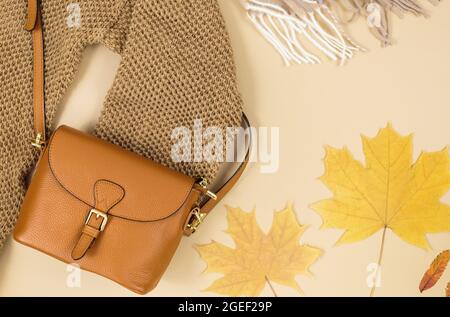 Orangefarbene Ledertasche, braune Schuhe, Schal, Herbstblätter auf grauem Hintergrund. Trendige Herbst-Accessoires. Koz Stockfoto