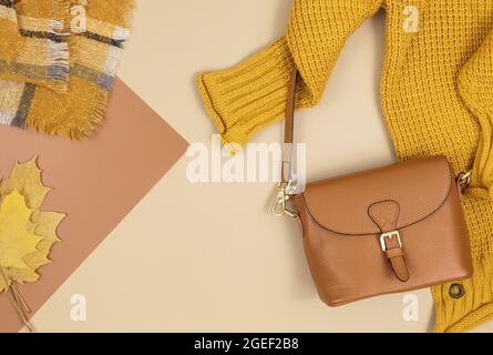 Orangefarbene Ledertasche, Strickpullover, Schal, Herbstblätter auf beigefarbenem braunen Hintergrund. Trendige Herbst-Accessoires. Gemütliches Outfit Stockfoto