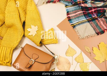 Orangefarbene Ledertasche, Strickpullover, Schal, Herbstblätter auf beigefarbenem braunen Hintergrund. Trendige Herbst-Accessoires. Gemütliches Outfit Stockfoto