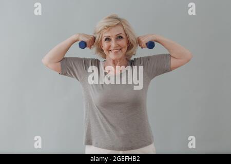 Ältere kaukasische alte Frau Porträt grauhaarig macht Übungen mit Hanteln zeigen Bizeps mit Lächeln auf grauem Hintergrund Stockfoto