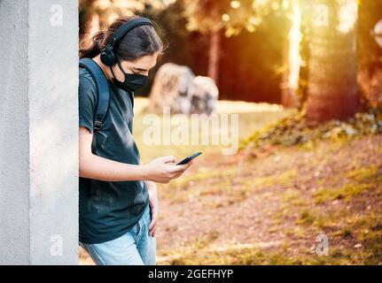 Teen High School Junge mit medizinischer Maske, Kopfhörer und Rucksack, SMS mit seinem Handy im Schulgarten. Stockfoto