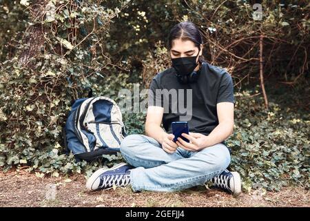 Teenager-High-School-Junge mit medizinischer Maske, Kopfhörern und Rucksack sitzt in gekreuzten Beinen im Schulgarten und SMS mit seinen Handys Stockfoto