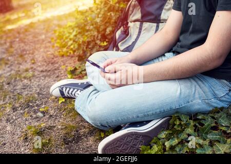 Junger Teenager-High-School-Junge, der im Schulgarten in gekreuzten Beinen sitzt und sein Mobiltelefon oder Smartphone benutzt Stockfoto