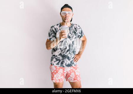 Im Sommer trinkt ein junger Mann mit Hemd, Mütze und Gläsern einen Cocktail mit Strohhalm, bei Tageslicht an einer weißen Wand Stockfoto