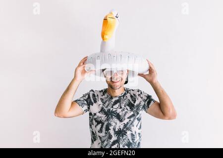 Junger Mann im Sommer mit Hemd, Mütze, Handtuch und Gläsern mit Blumenmuster, mit einer Ente auf dem Kopf, bei Tageslicht auf einer weißen Wand Stockfoto