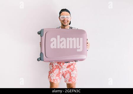 Junger Mann in einem Hemd mit Blumenmuster und Mütze, mit einem Koffer, um in den Urlaub zu gehen, bei Tageslicht auf einer weißen Wand Stockfoto