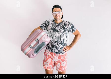 Junger Mann in einem Hemd mit Blumenmuster und Mütze, mit einem Koffer, um in den Urlaub zu gehen, bei Tageslicht auf einer weißen Wand Stockfoto