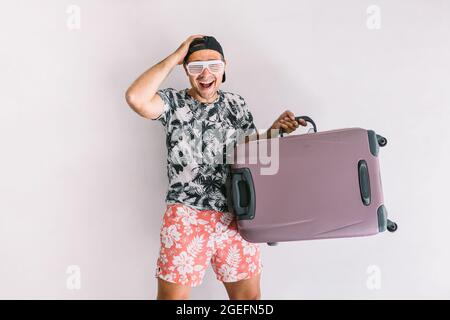 Junger Mann in einem Hemd mit Blumenmuster und Mütze, mit einem Koffer, um in den Urlaub zu gehen, bei Tageslicht auf einer weißen Wand Stockfoto