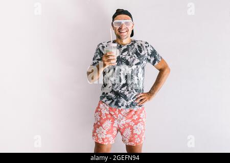 Im Sommer trinkt ein junger Mann mit Hemd, Mütze und Gläsern einen Cocktail mit Strohhalm, bei Tageslicht an einer weißen Wand Stockfoto