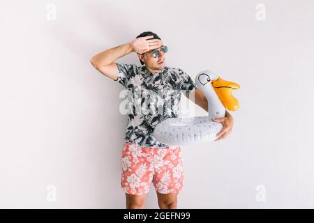 Junger Mann mit geblümtem Hemd, Mütze und Brille im Sommer mit einem Entlein, das Wärme Gesten macht, bei Tageslicht auf einer weißen Wand Stockfoto