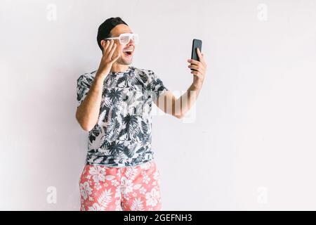 Im Sommer macht ein junger Mann mit Blumenhemd, Mütze und Brille ein Selfie und eine Videokonferenz mit seinem Smartphone und trinkt einen Cocktail mit einem Strohhalm Stockfoto