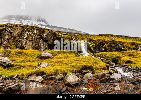 Schneebedeckte Berge und Wasserfälle am Strand am Reydarfjordur Fjord, Island Stockfoto