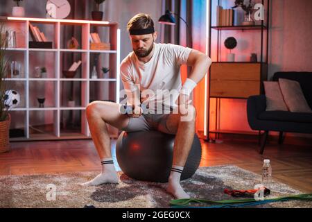Gesundes Lifestyle-Konzept. Vorderansicht eines hübschen, aktiven jungen bärtigen Mannes im Alter von 30 Jahren in Sportkleidung, der zu Hause auf einem Fitball sitzt und mit Hanteln Übungen macht, um seinen Körper gesund und stark zu halten. Stockfoto