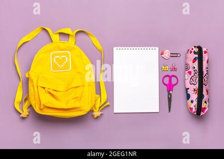 Zurück zur Schule, Bildungskonzept. Gelber Rucksack mit Schulbedarf - Notizbuch, Stifte, Clips, Schere isoliert auf violettem Hintergrund Draufsicht Copy Stockfoto