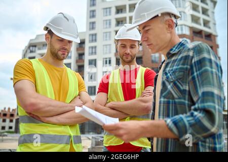 Drei kaukasische Arbeiter in Kopfbedeckung, die technische Zeichnungen analysierten Stockfoto