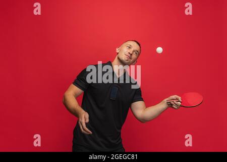 Ein junger kaukasischer Mann mit Tischtennisschläger, der Tischtennis auf rotem Hintergrund spielt. Stockfoto