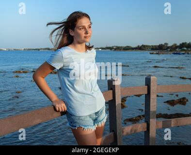 Teen Mädchen steht auf einer Holzbrücke am Meer bei Sonnenuntergang. Teenager mit blauem T-Shirt und Jeans-Shorts, die zur Seite schauen, drei Viertel lang geschossen. Stockfoto